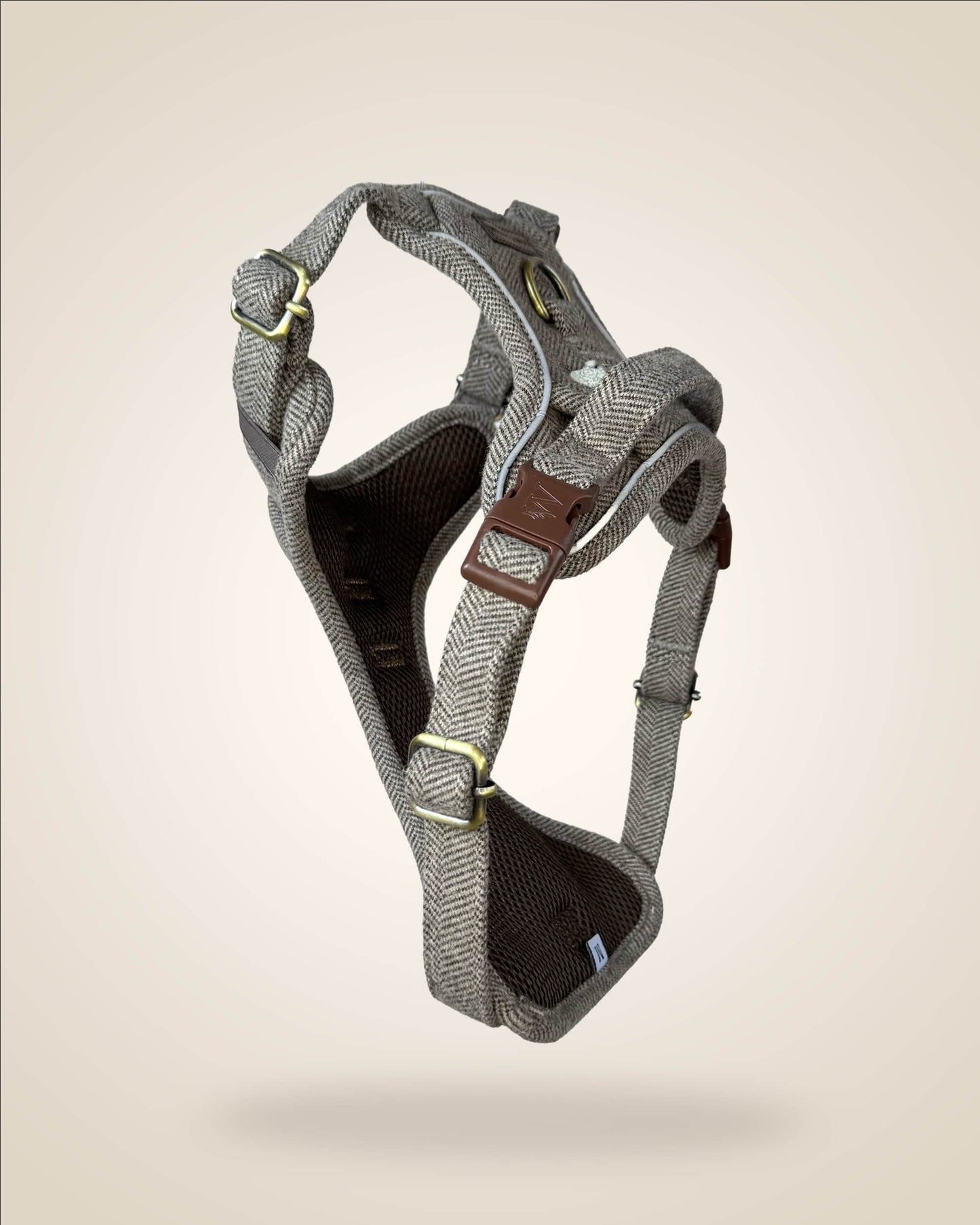 A brown and white tweed herringbone patterned dog harness with white sheep motifs, bronze tone hardware, and a brown leather patch, shown against a light beige background.