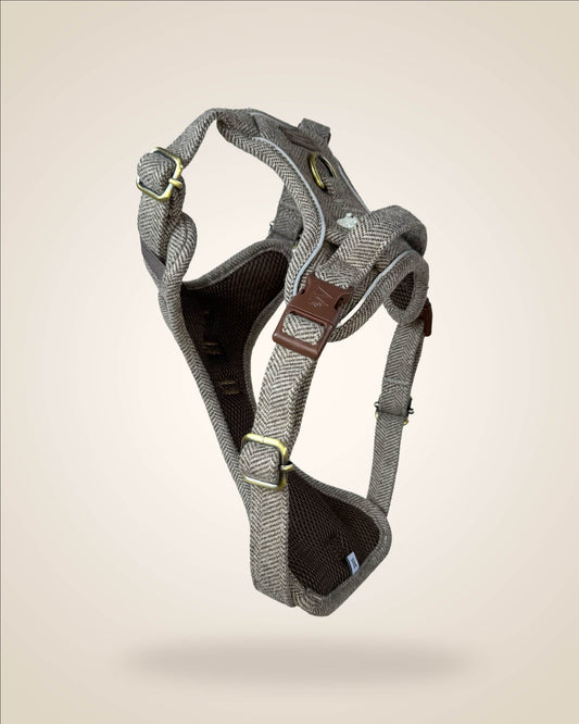 A brown and white tweed herringbone patterned dog harness with white sheep motifs, bronze tone hardware, and a brown leather patch, shown against a light beige background.