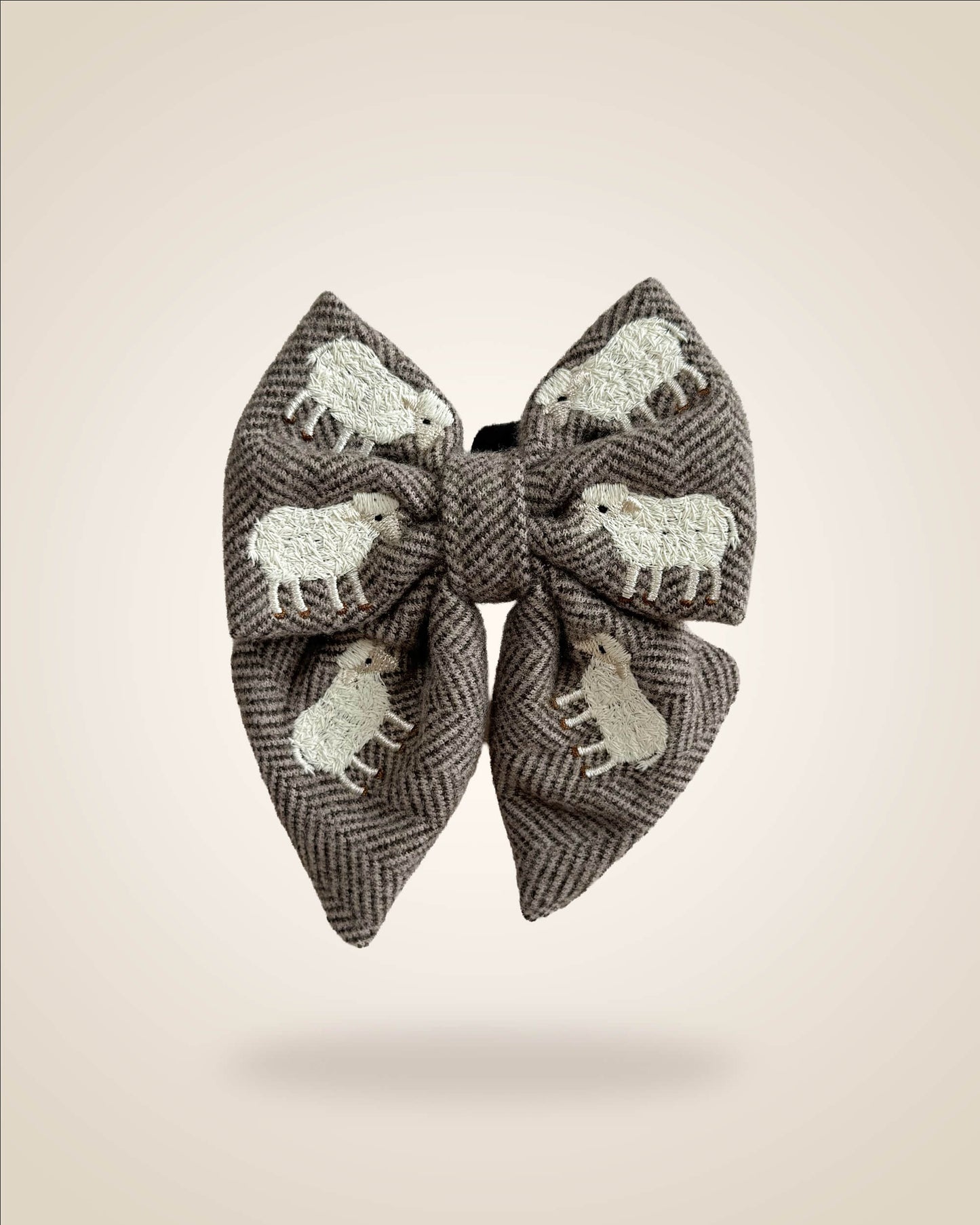 A large fabric bow in a brown and gray herringbone pattern, decorated with white embroidered sheep motifs, shown against a solid beige background.