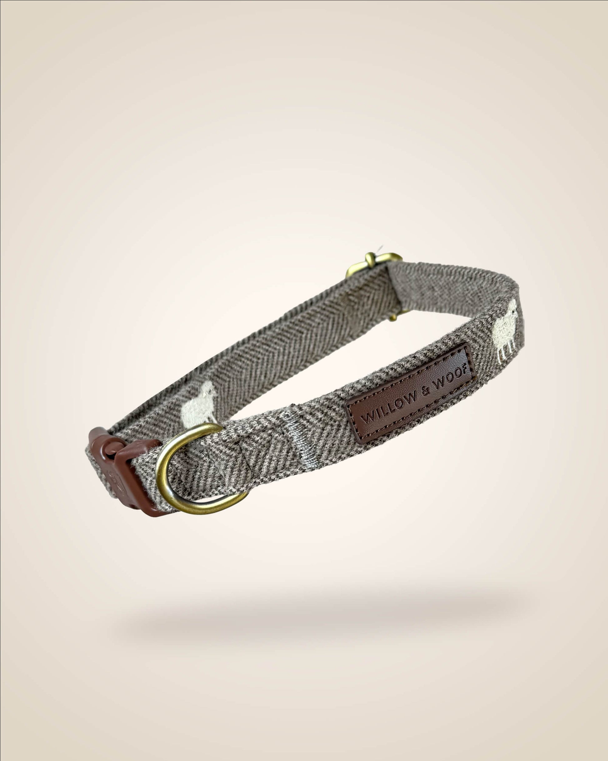 A brown and white tweed herringbone patterned dog collar with white sheep motifs, bronze tone hardware, and a brown leather patch, shown against a light beige background.