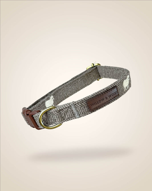 A brown and white tweed herringbone patterned dog collar with white sheep motifs, bronze tone hardware, and a brown leather patch, shown against a light beige background.