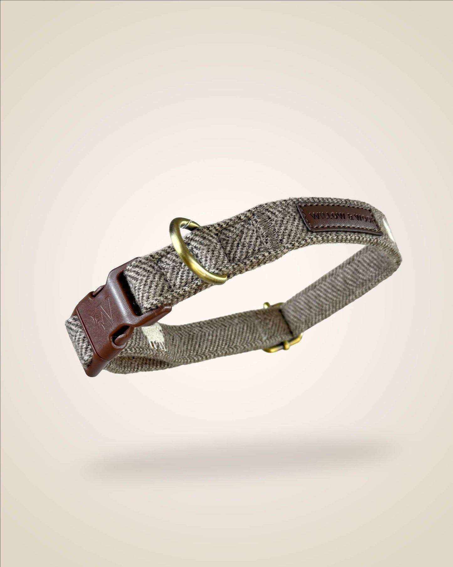 A brown and white tweed herringbone patterned dog collar with white sheep motifs, bronze tone hardware, and a brown leather patch, shown against a light beige background.