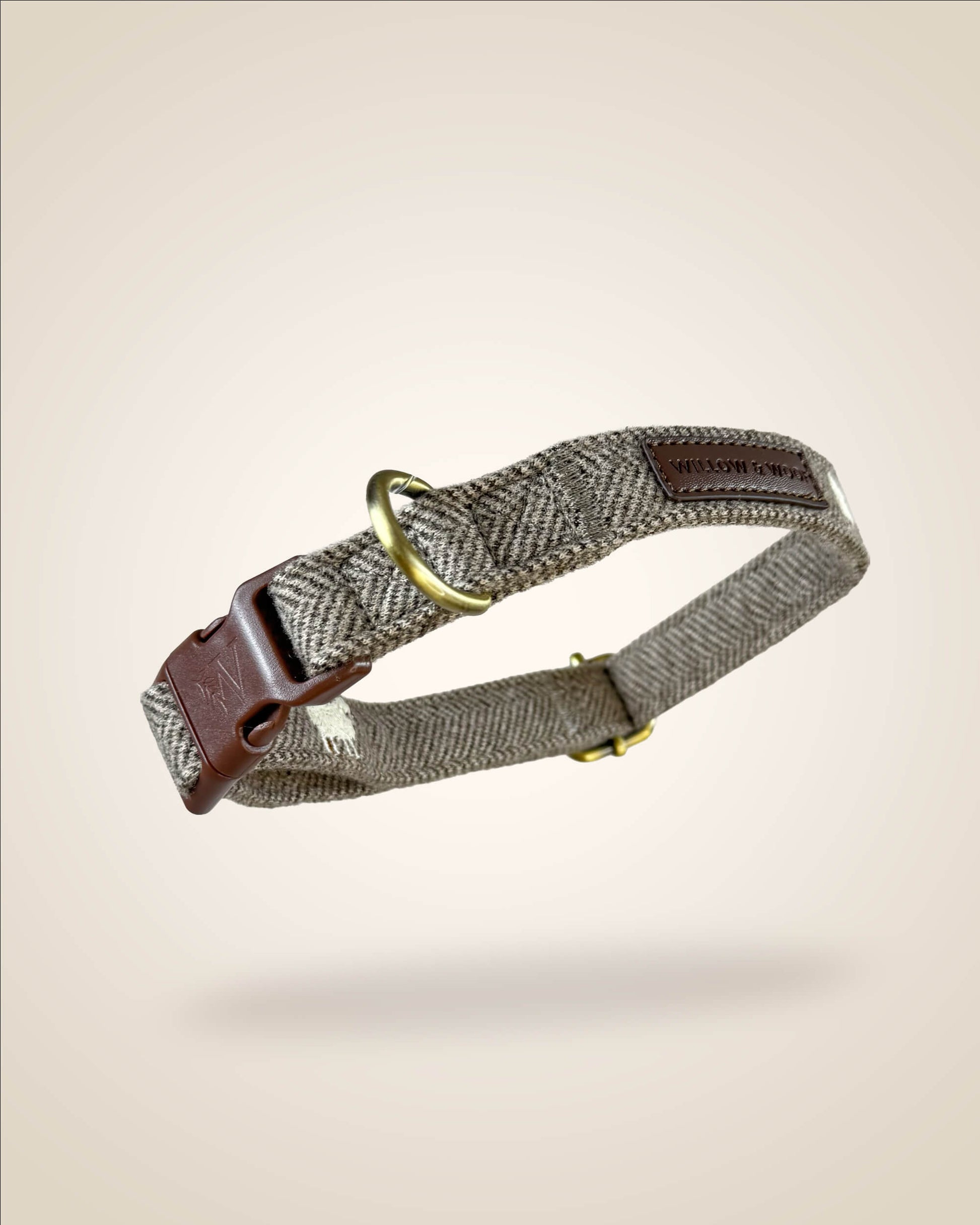 A brown and white tweed herringbone patterned dog collar with white sheep motifs, bronze tone hardware, and a brown leather patch, shown against a light beige background.