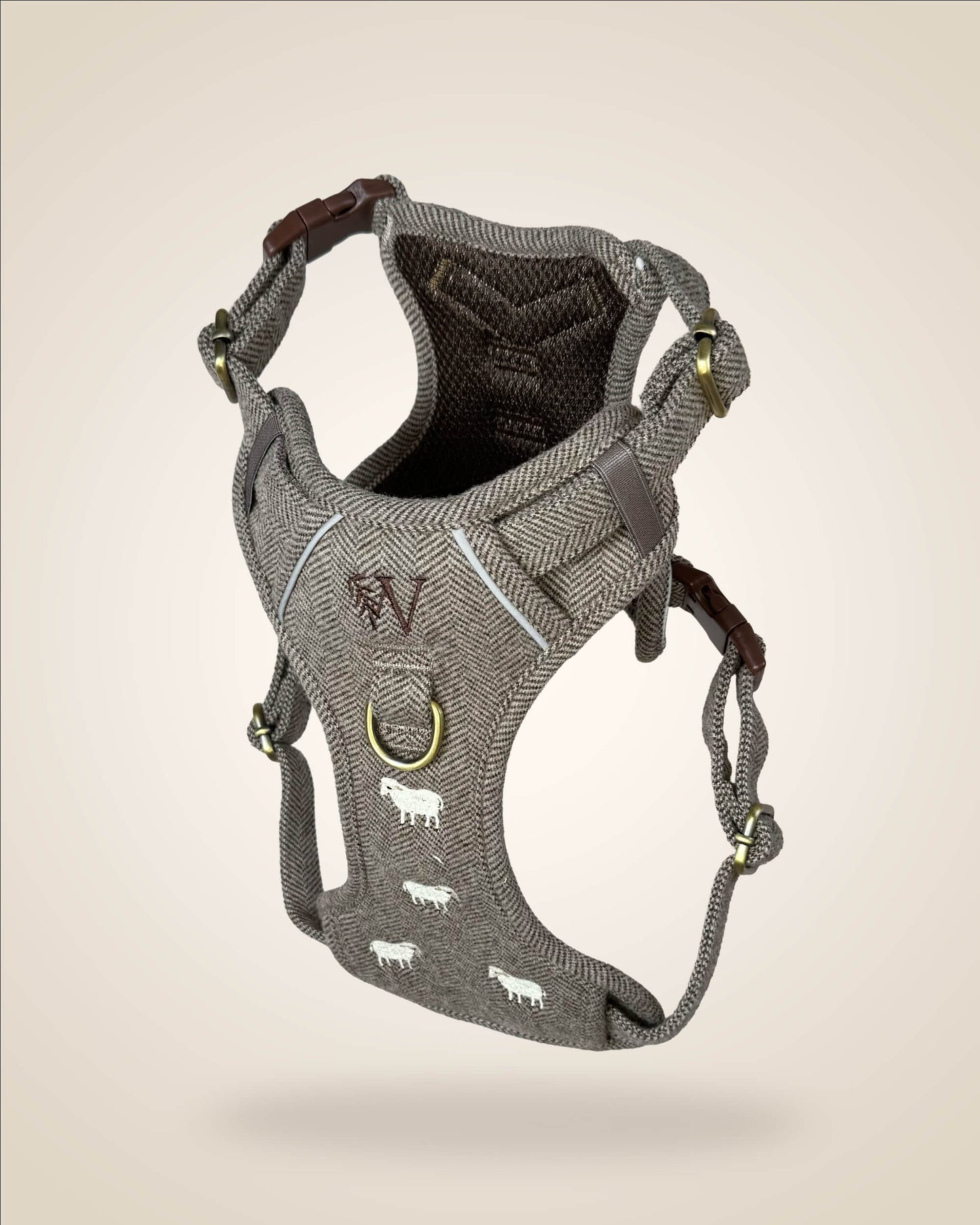 A brown and white tweed herringbone patterned dog harness with white sheep motifs, bronze tone hardware, and a brown leather patch, shown against a light beige background.