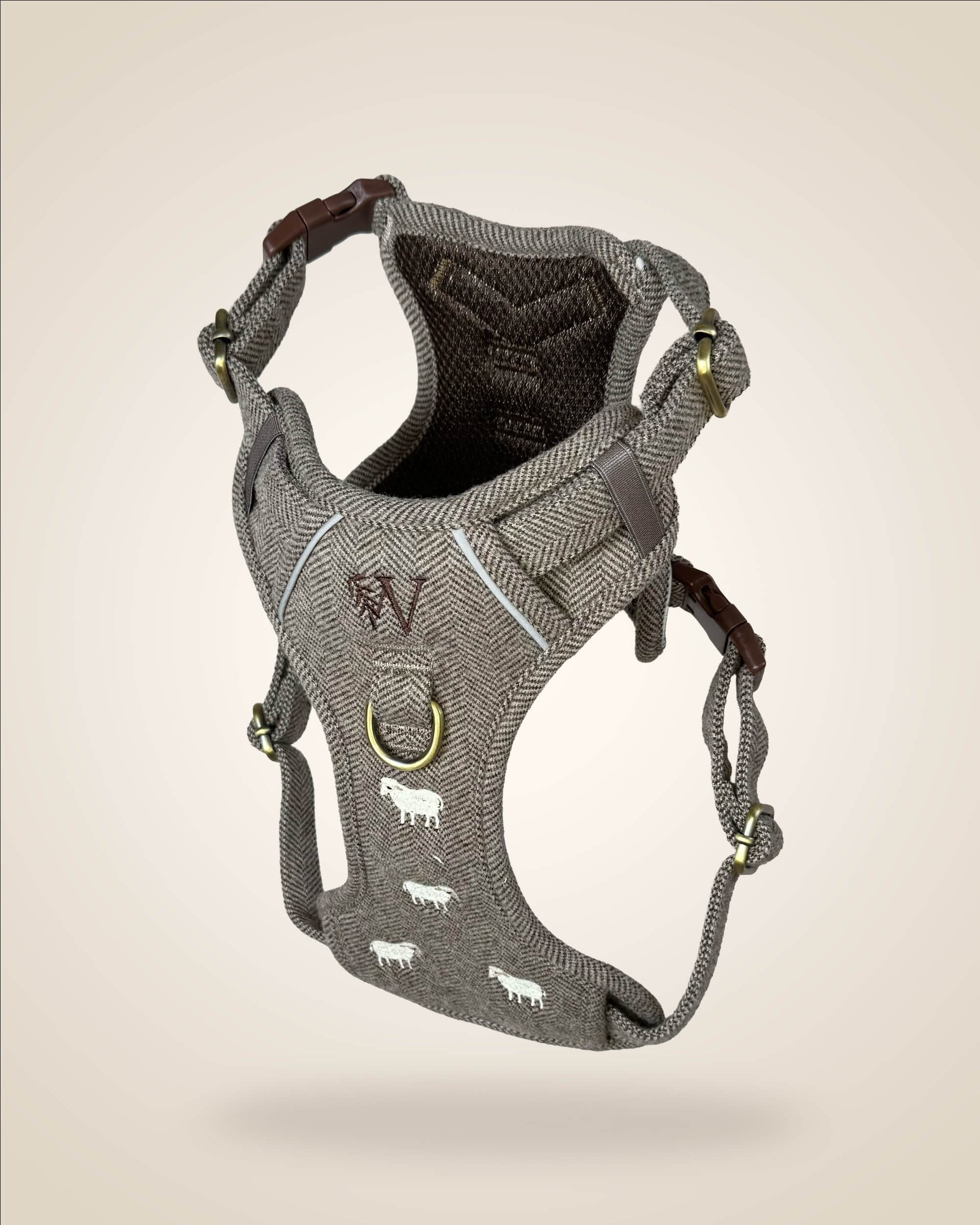 A brown and white tweed herringbone patterned dog harness with white sheep motifs, bronze tone hardware, and a brown leather patch, shown against a light beige background.