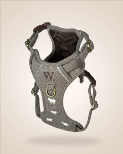 A brown and white tweed herringbone patterned dog harness with white sheep motifs, bronze tone hardware, and a brown leather patch, shown against a light beige background.