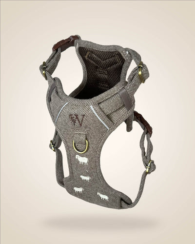 A brown and white tweed herringbone patterned dog harness with white sheep motifs, bronze tone hardware, and a brown leather patch, shown against a light beige background.