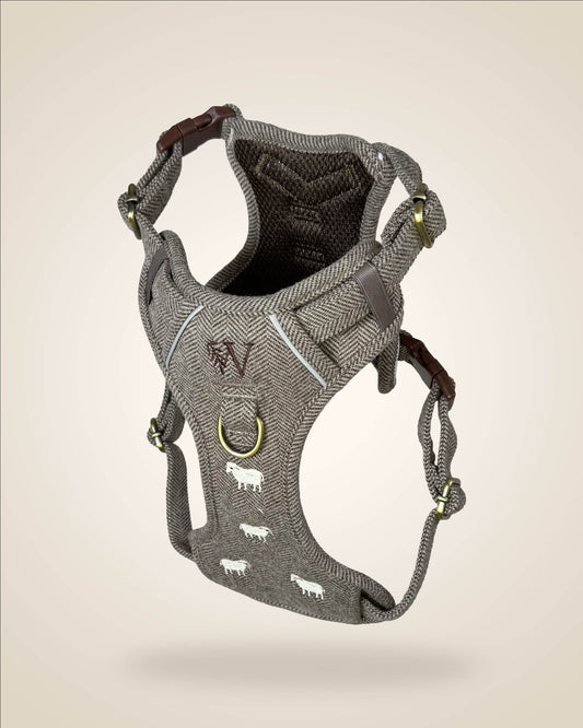 A brown and white tweed herringbone patterned dog harness with white sheep motifs, bronze tone hardware, and a brown leather patch, shown against a light beige background.