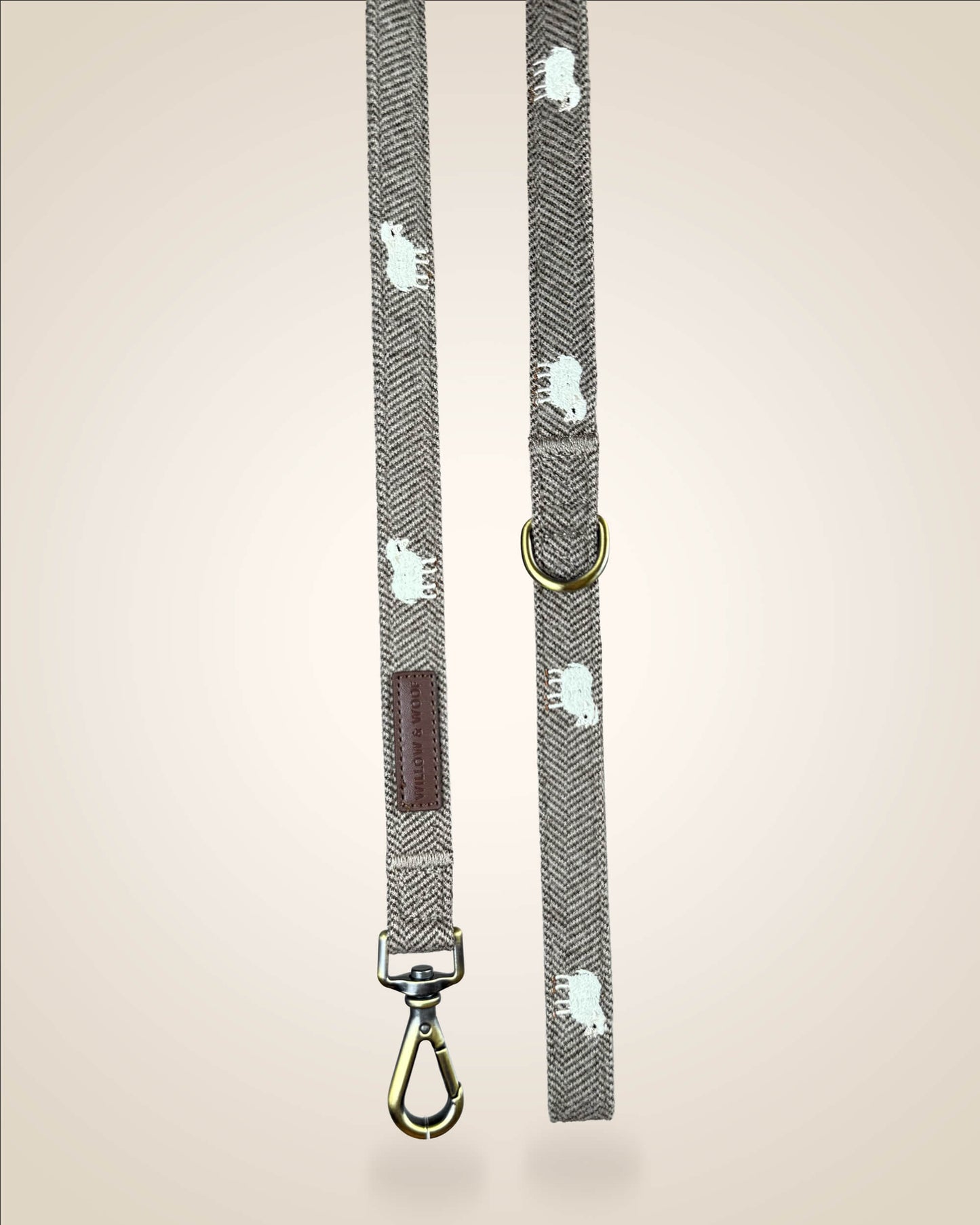 A brown and white tweed herringbone patterned dog lead with white sheep motifs, bronze tone hardware, and a brown leather patch, shown against a light beige background.