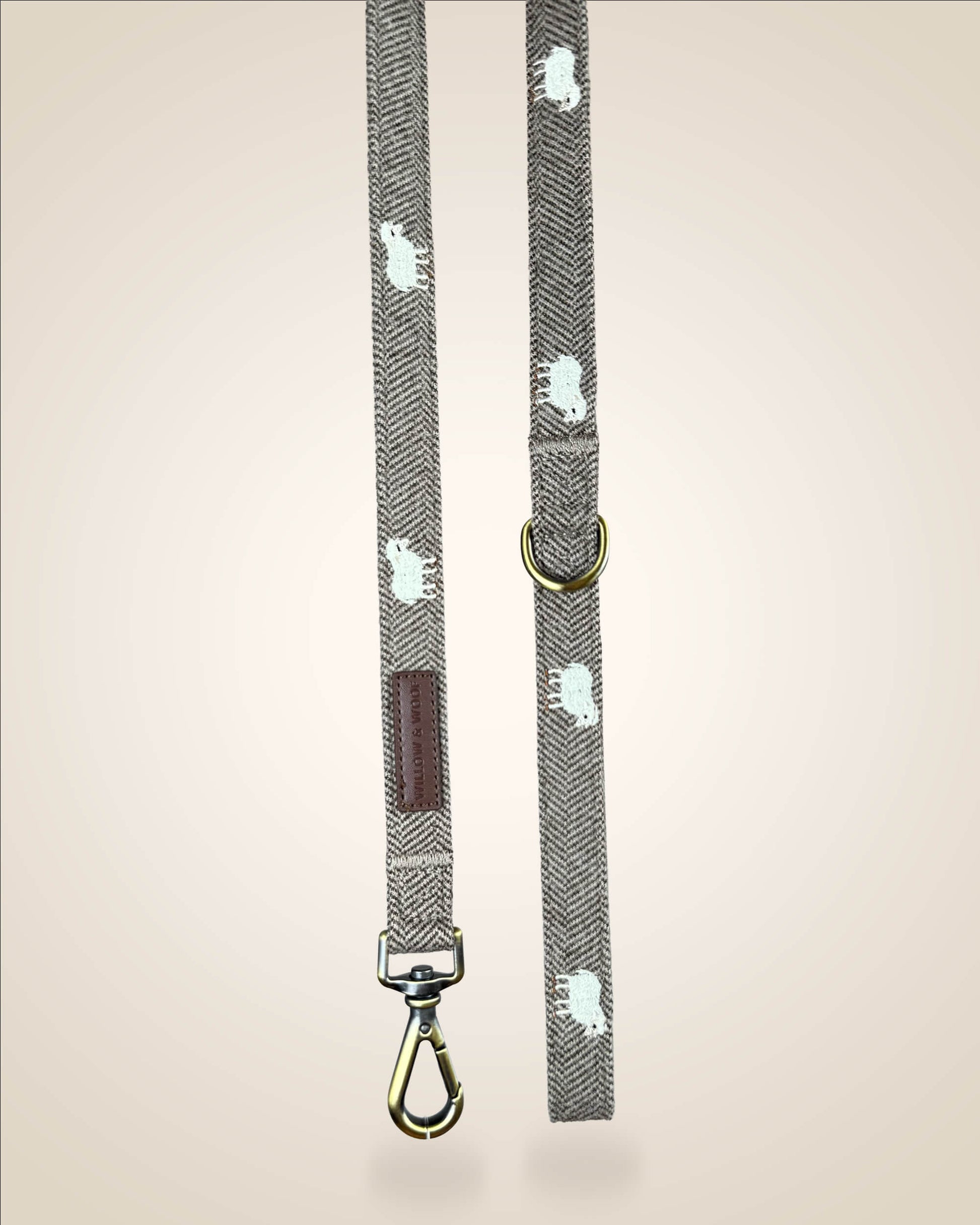 A brown and white tweed herringbone patterned dog lead with white sheep motifs, bronze tone hardware, and a brown leather patch, shown against a light beige background.