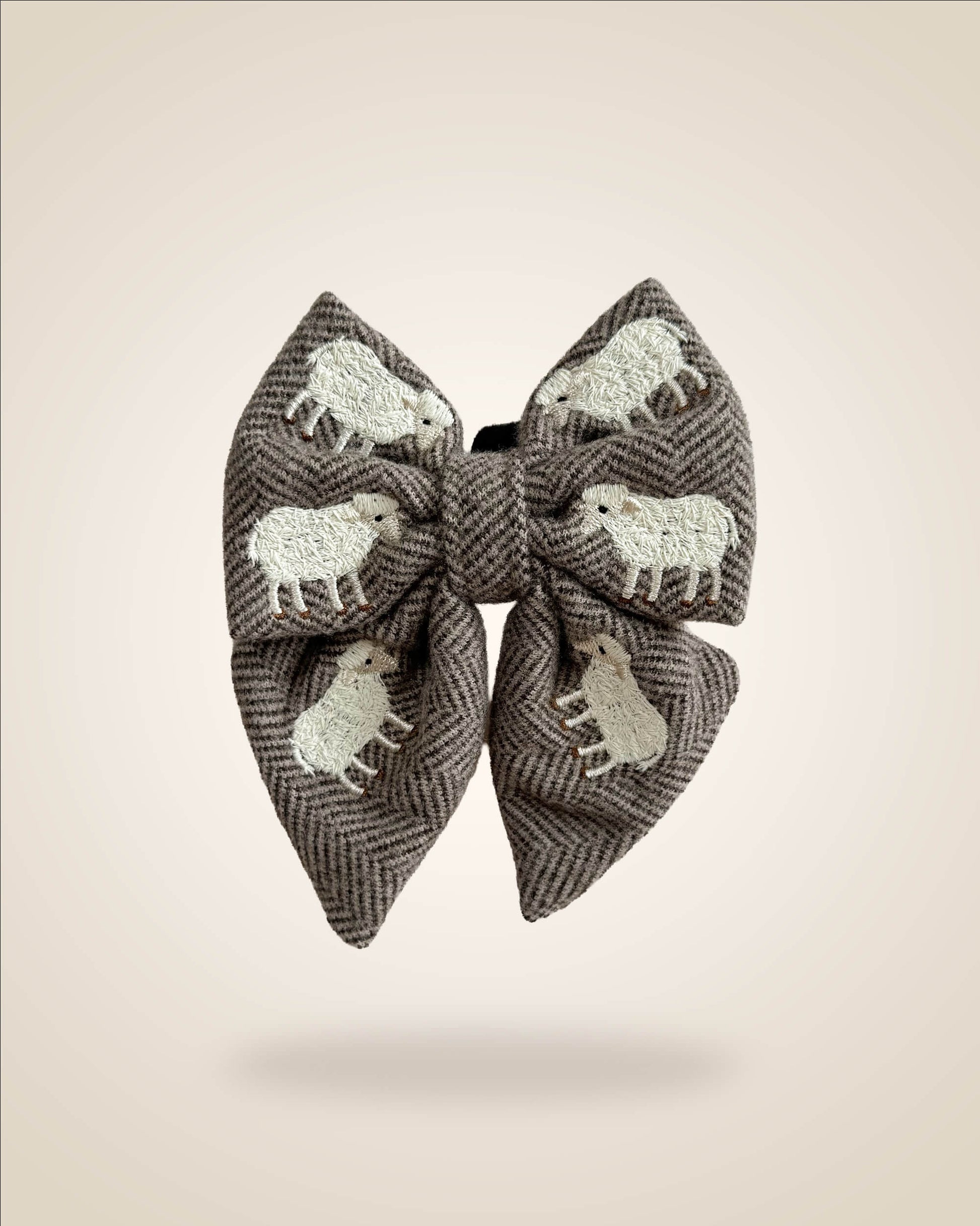 A large fabric bow in a brown and gray herringbone pattern, decorated with white embroidered sheep motifs, shown against a solid beige background.
