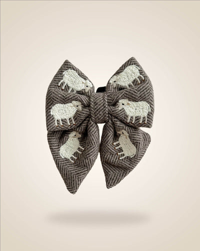 A large fabric bow in a brown and gray herringbone pattern, decorated with white embroidered sheep motifs, shown against a solid beige background.