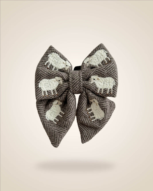 A large fabric bow in a brown and gray herringbone pattern, decorated with white embroidered sheep motifs, shown against a solid beige background.
