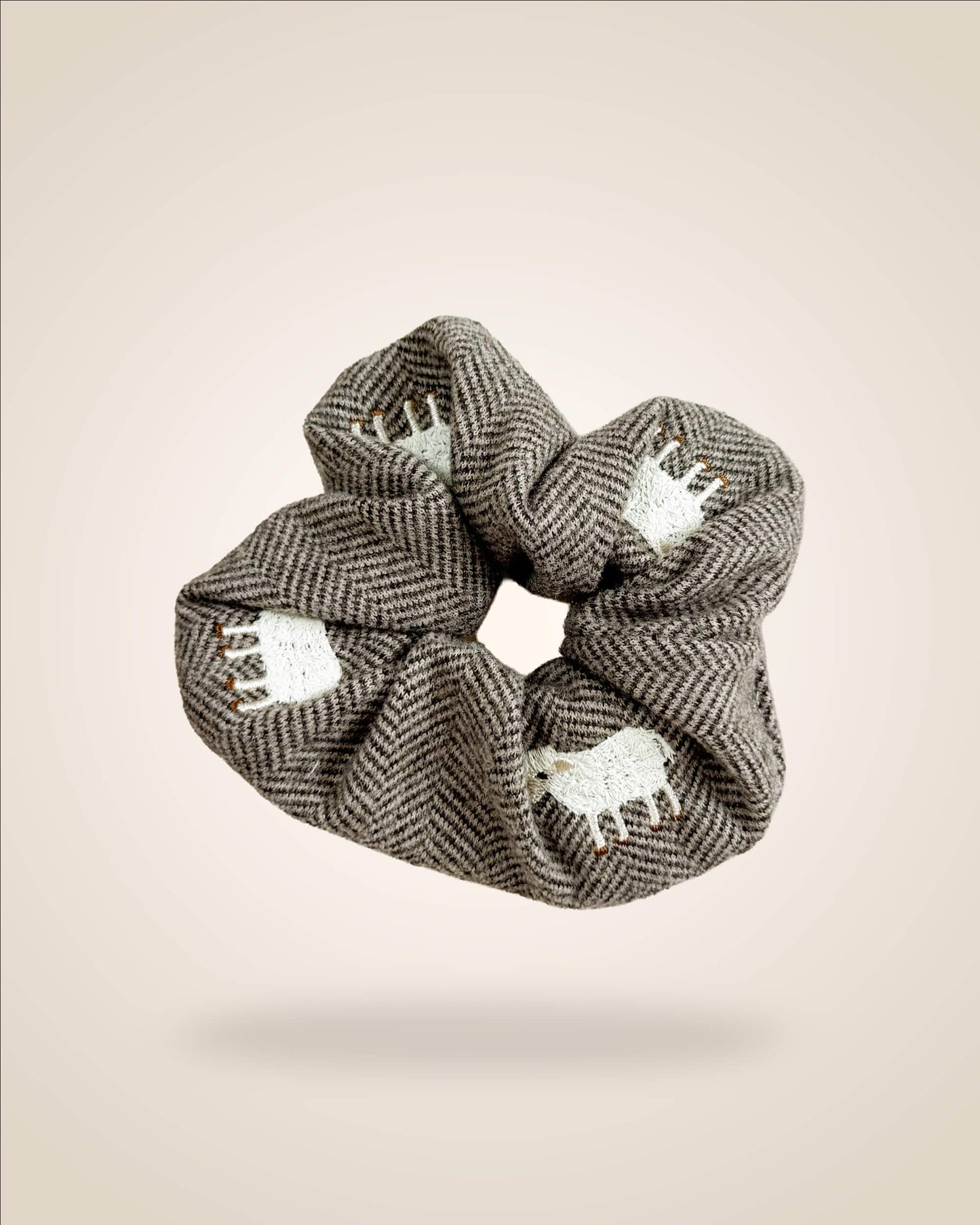 A fabric hair scrunchie featuring a brown and gray herringbone pattern with white embroidered sheep motifs, shown floating against a solid beige background.