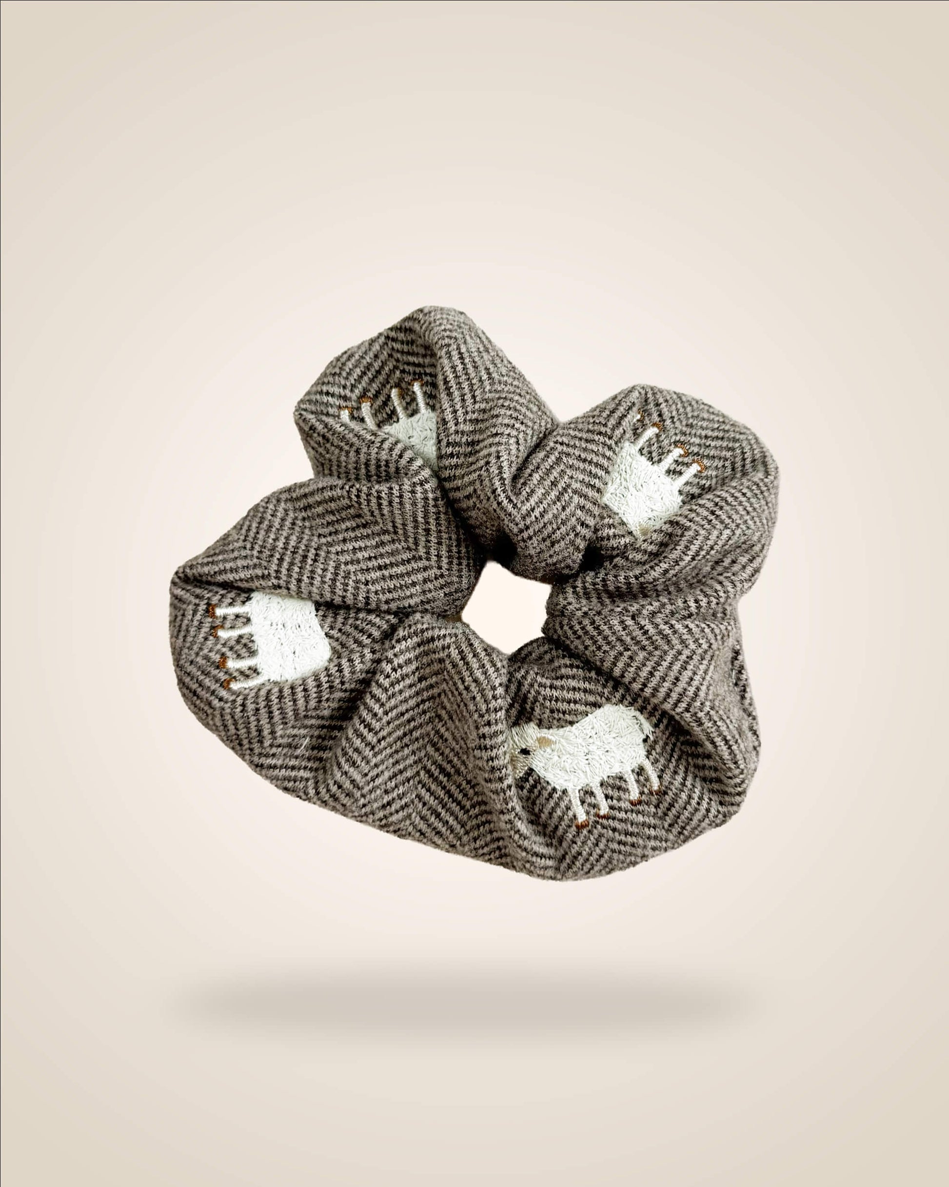 A fabric hair scrunchie featuring a brown and gray herringbone pattern with white embroidered sheep motifs, shown floating against a solid beige background.