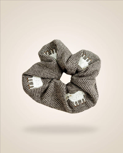 A fabric hair scrunchie featuring a brown and gray herringbone pattern with white embroidered sheep motifs, shown floating against a solid beige background.